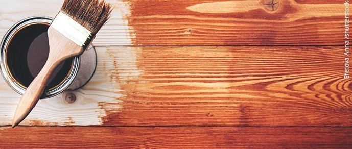 Eine geöffnete Dose Glasur neben einem Pinsel, der auf einer teilweise bemalten Holzoberfläche liegt.